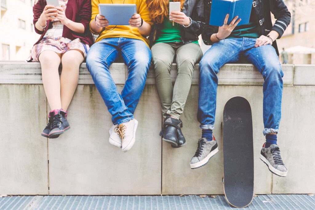 Four teenagers shown from mid shoulders down sat on a concrete wall. Two are looking at phones, one is looking at a tablet and another is reading a book. A skateboard is propped against the wall.