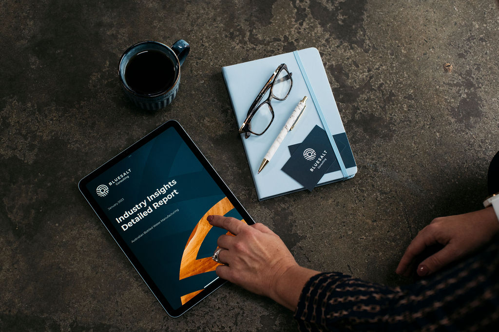 BlueSalt Consulting Industry Insights Report, an example of quality research, on an ipad, with woman's hand pointing at it, sat next to a mug, notebook, reading glasses, pen and business cards.