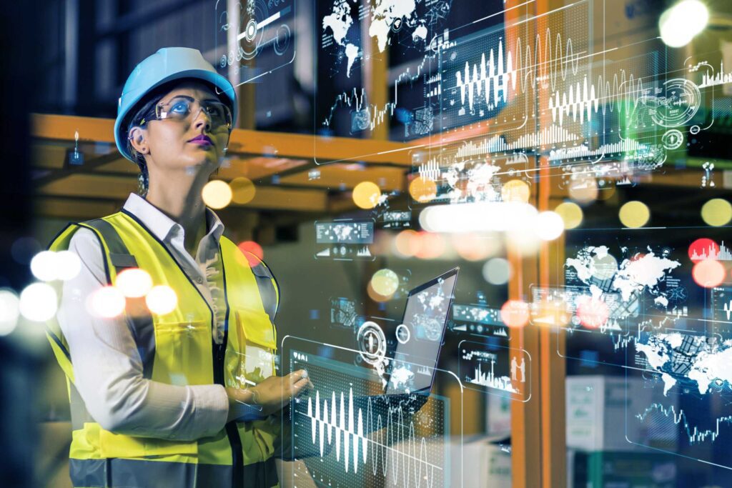 Woman wearing hard hat, safety glass and high vis jacket operating high tech equipment in factory setting.